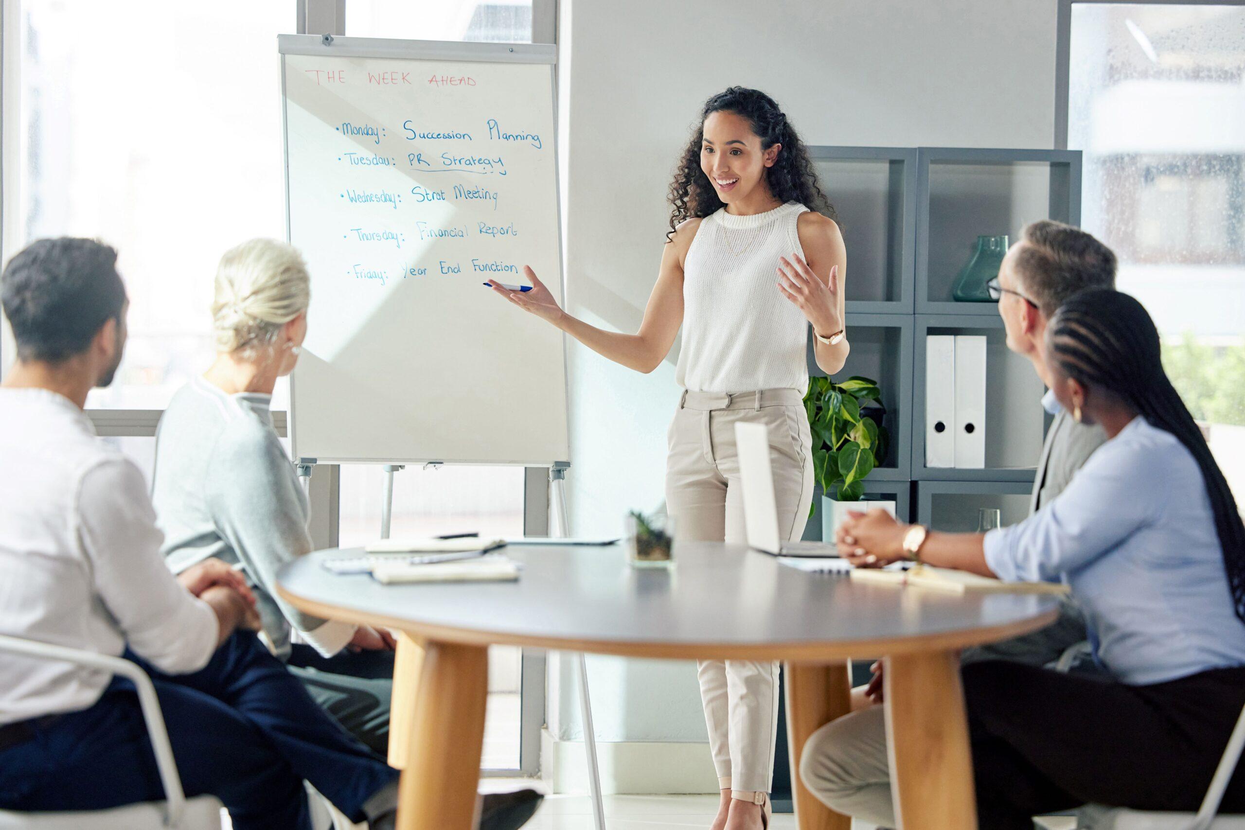 Mehrere Personen haben ein Meeting. Eine Frau steht vorne und ist am reden  