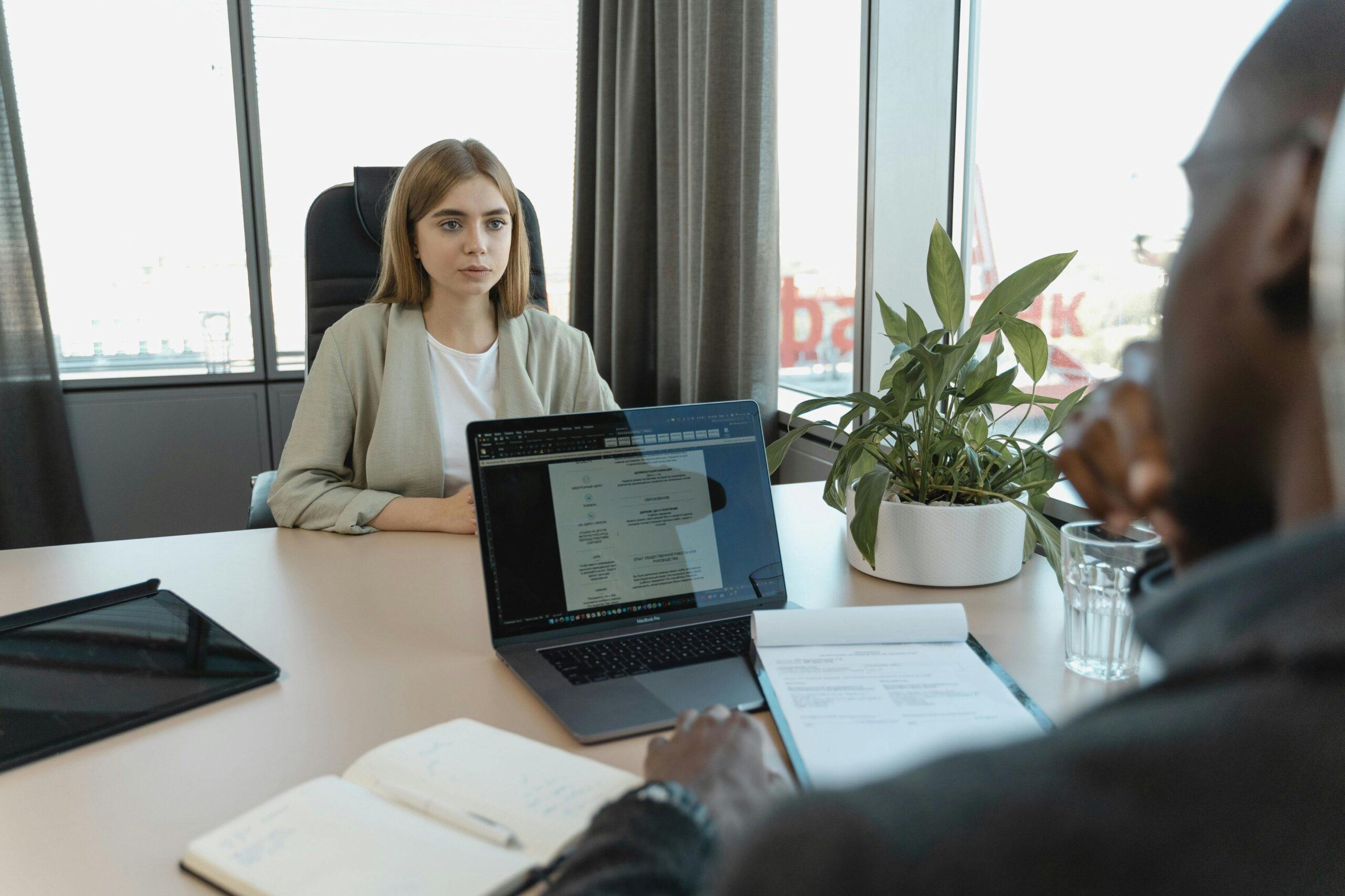 Eine Frau hat ein Vorstellungsgespräch und sitzt in einem Büro von einem Mann. Sie Sprechen miteinander und der Mann hat einen Laptop vor sich stehebn