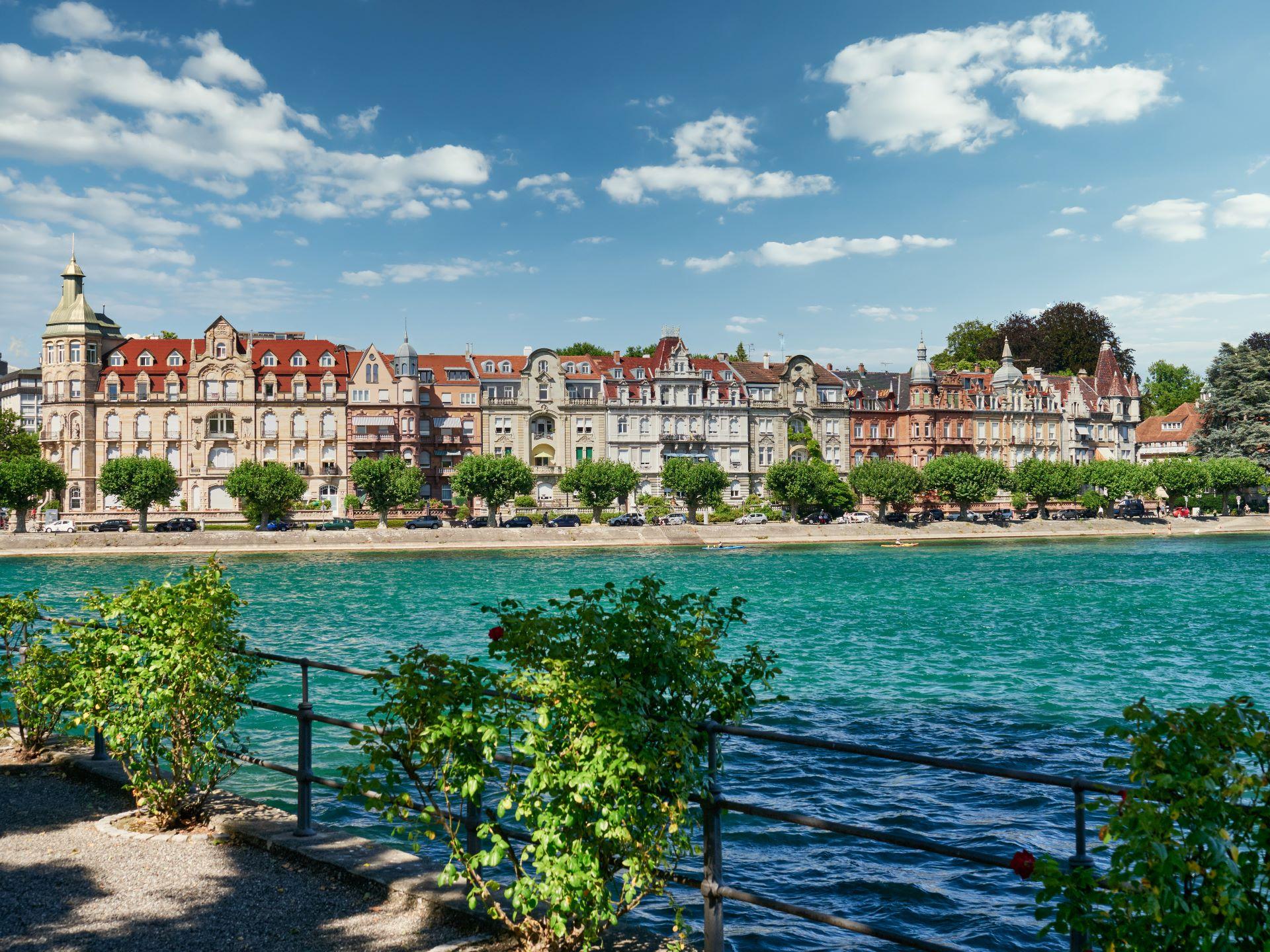 Malerische Uferpromenade am Bodensee mit historischen Gebäuden und klarem, blauem Wasser unter einem sonnigen Himmel.