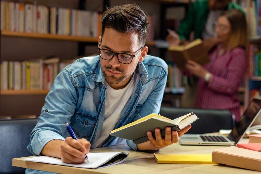 Junger männlicher Student lernt in der Bibliothek und liest Buch.